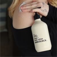 A person applying white body lotion onto their shoulder. The bottle is a warm cream color with black text and a black pump.
