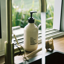 A bottle of Rose Crush Amber body wash sits on a kitchen sink counter with running water, a brass faucet, and a wooden scrub brush nearby. The bottle is cream-colored with a black pump and black text.